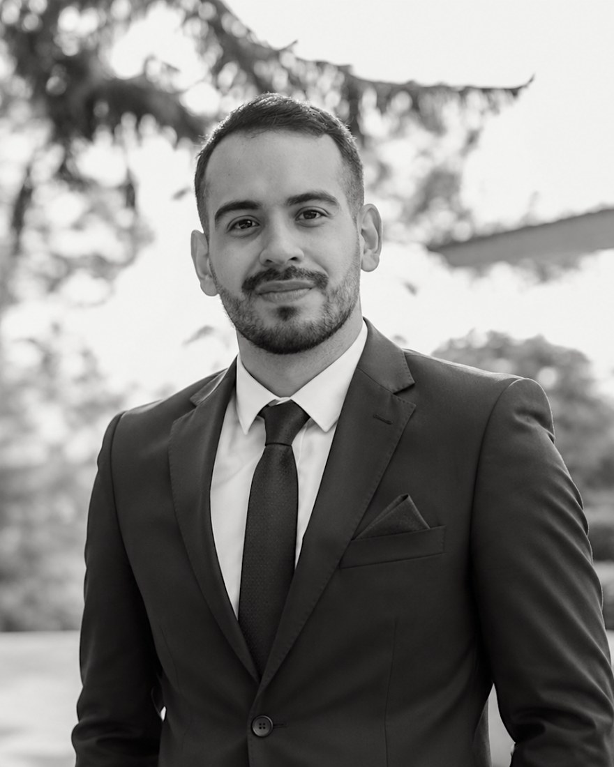 David Monzón, independent AI systems consultant, in a dark suit and tie against a blurred outdoor background, in black and white.